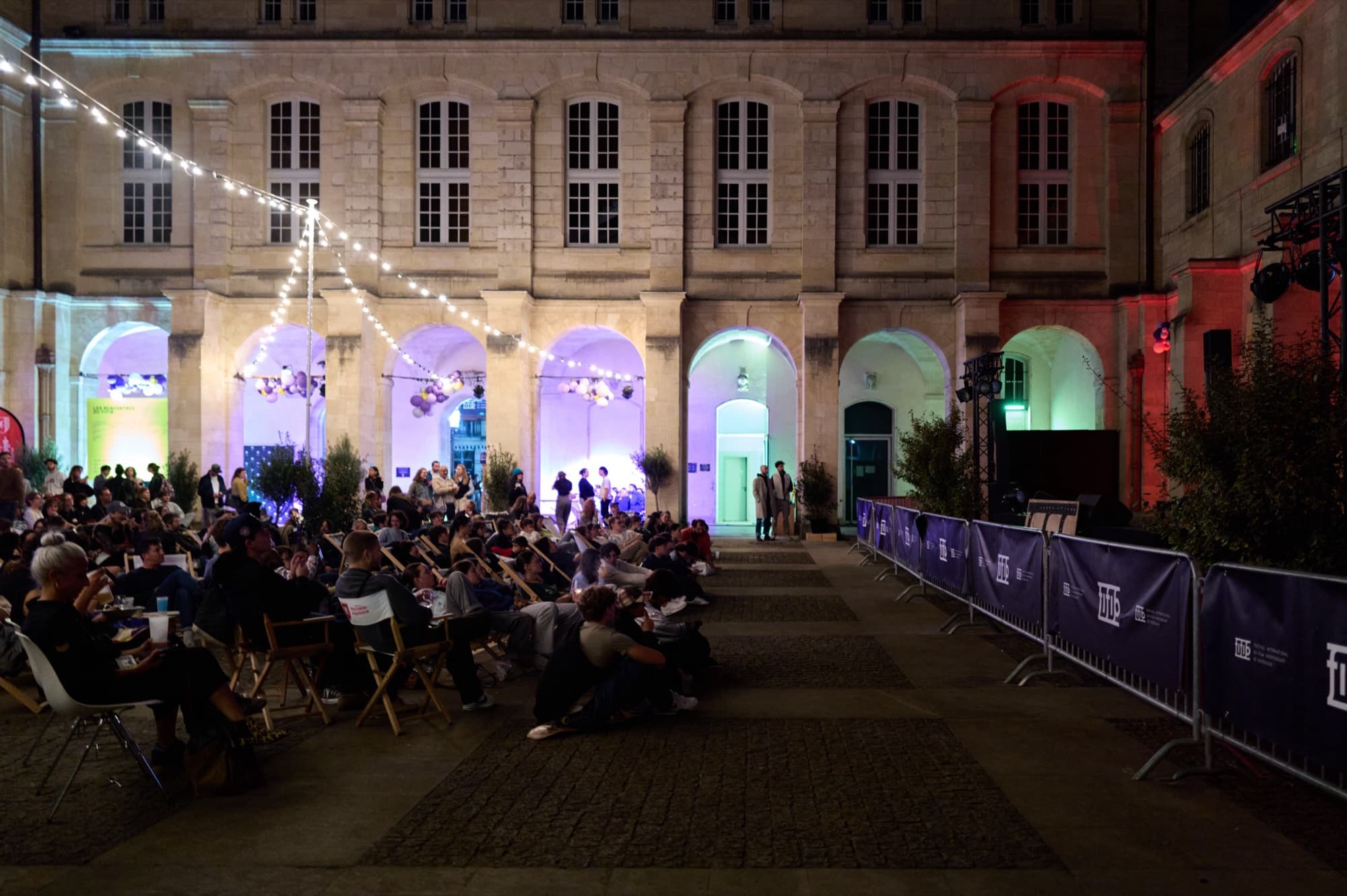 Public assis dans la cour Mably pendant une soirée du Village du FIFIB, façade éclairée en bleu